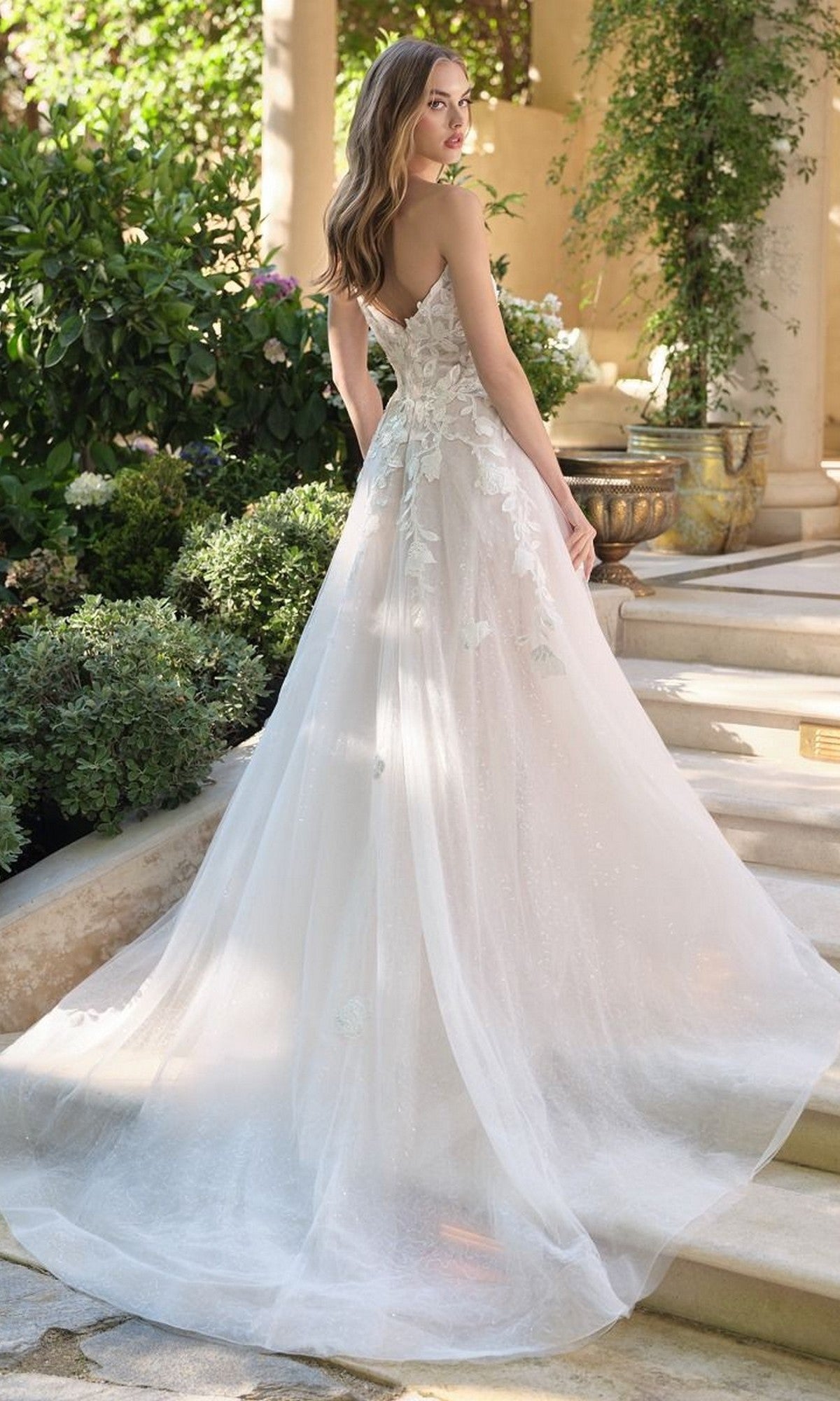 A woman wearing the Andrea and Leo WL070 long wedding dress, featuring a white strapless bodice with 3D floral lace appliques and an a-line tulle skirt, stands on garden stone steps, glancing over her shoulder.